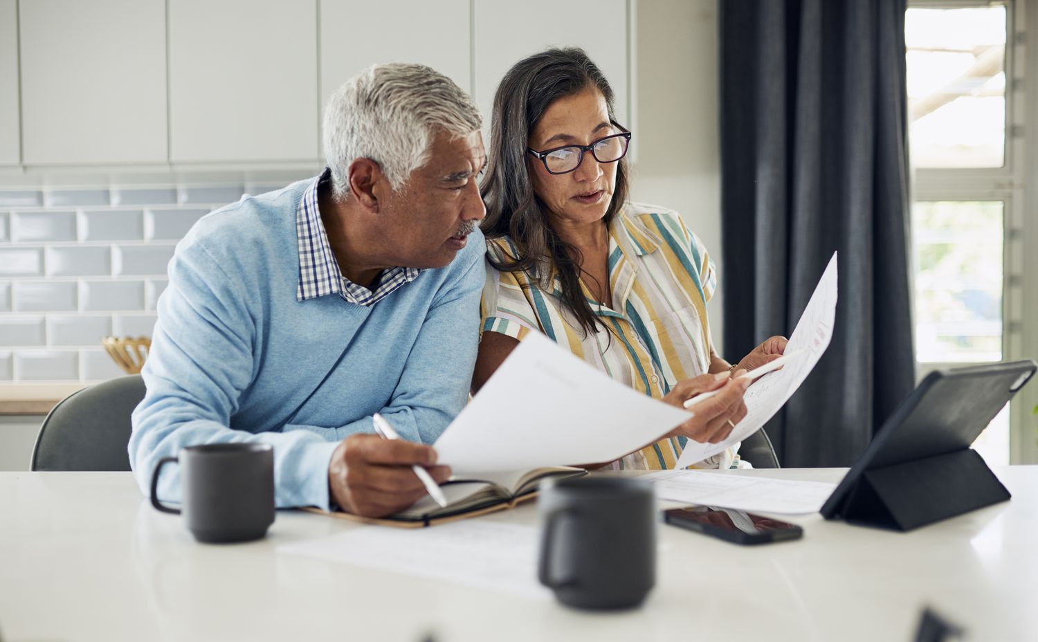 The image shows an older couple sitting together at a table, reviewing documents. The man, wearing a light blue sweater over a checkered shirt, looks intently at the papers, while the woman, in a striped shirt and glasses, appears to explain or discuss the details. The table holds an open notebook, a tablet on a stand, and a few mugs. The background features a modern kitchen with white tiled walls and natural light coming through a window. The scene suggests a collaborative effort, possibly related to finances or planning.