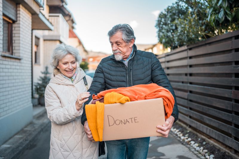 A man and a woman walk down a street carrying a box that has clothes in it and says "donate" on the side