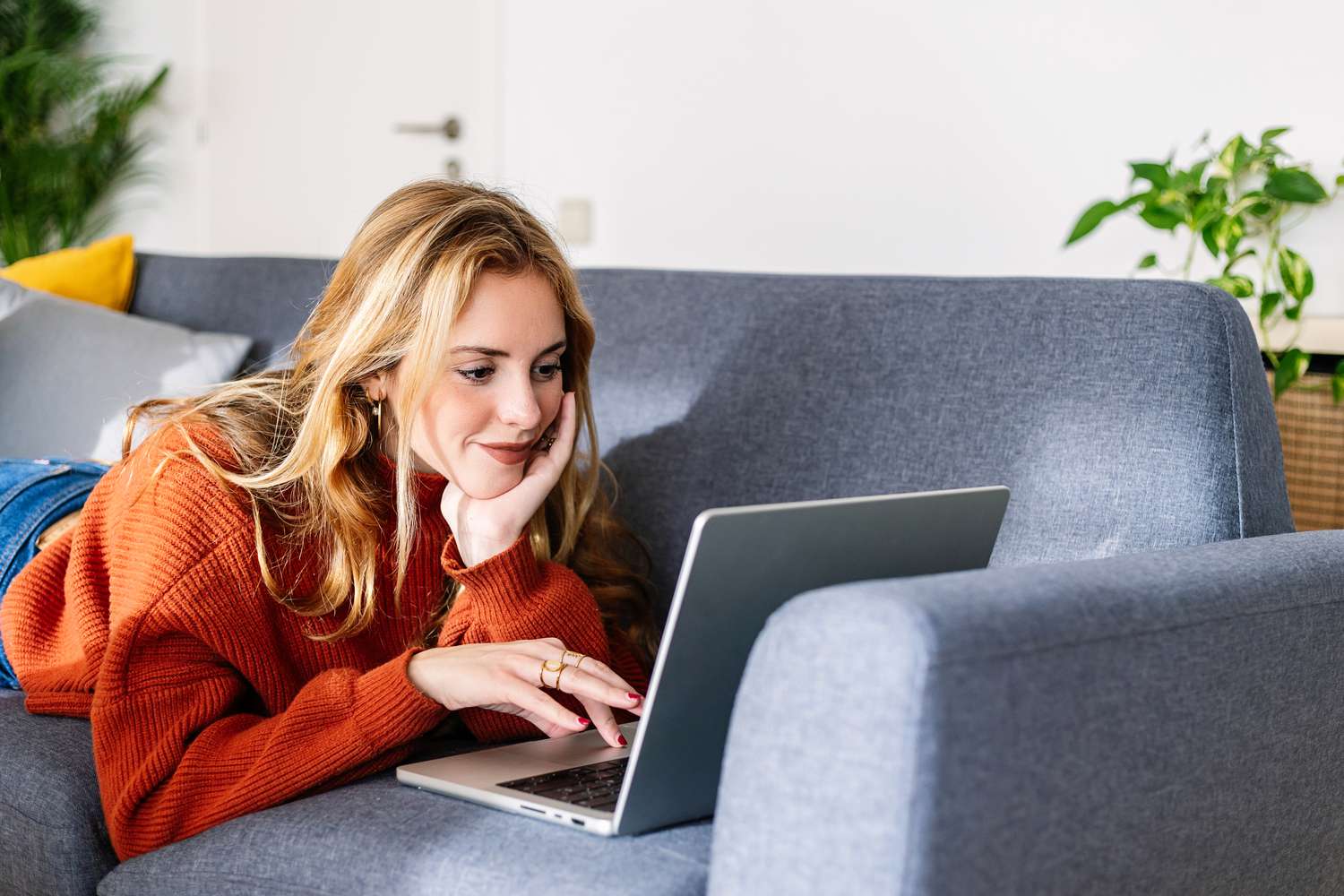 A woman in an orange sweater lays on her couch looking at a laptop