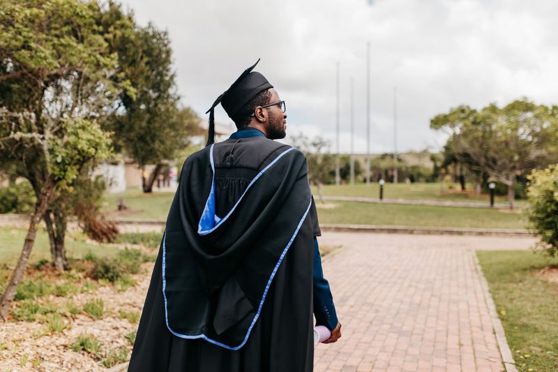 Young male graduate with beard wearing graduation gown and mortarboard walking and looking sideways into distance on campus grounds
