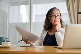 A person holds papers and looks at a laptop screen