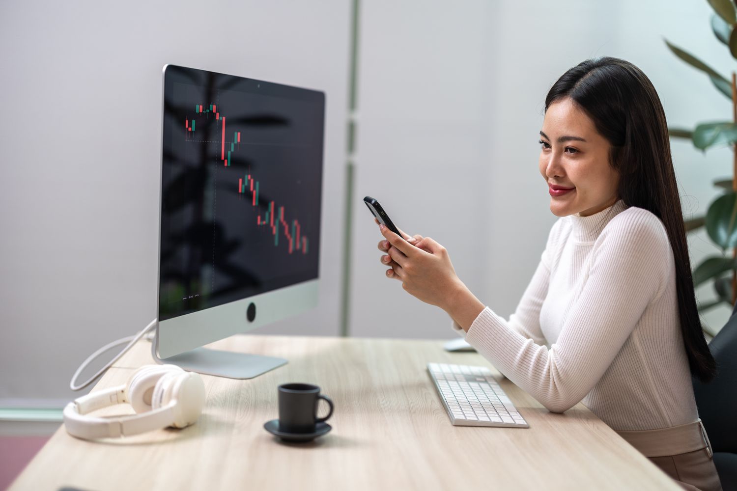 Woman sitting at a desk looking at a chart on a desk monitor while holding a cell phone.