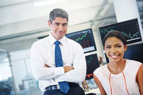 Two positive-looking financial officers in front of monitors displaying stock information.