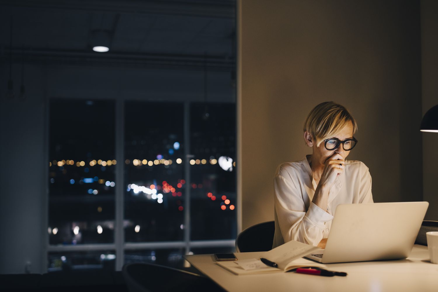 Stressed-looking mature businesswoman using laptop while sitting at illuminated desk in coworking space