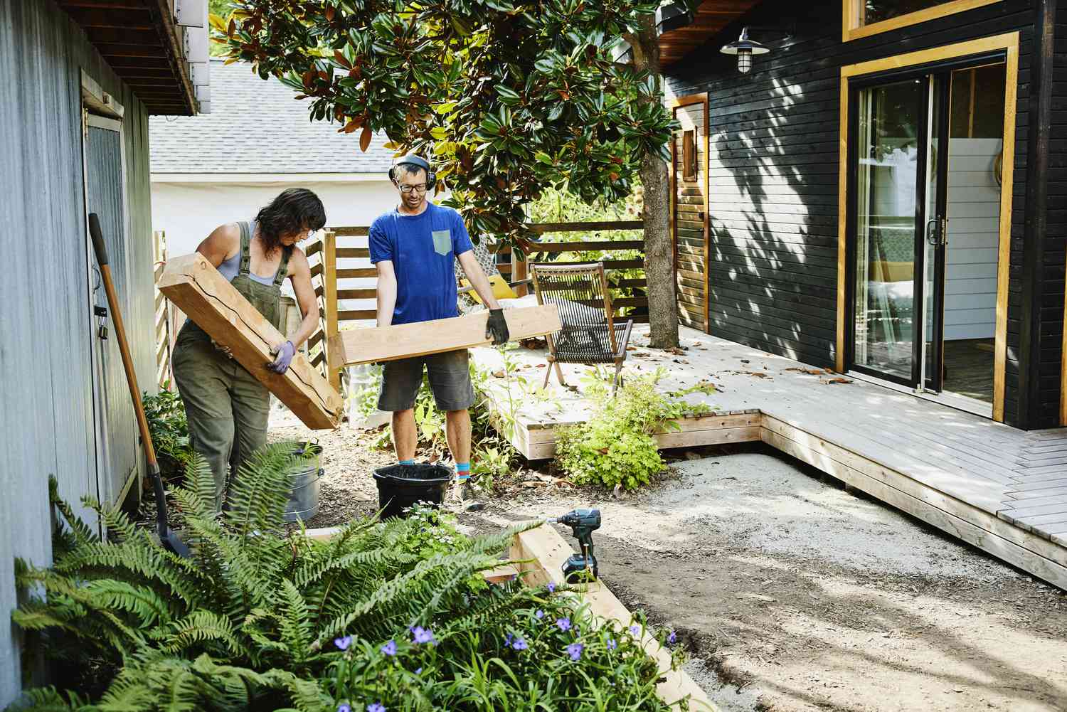 A couple carrying lumber works on raised flowerbeds on the exterior of their home.