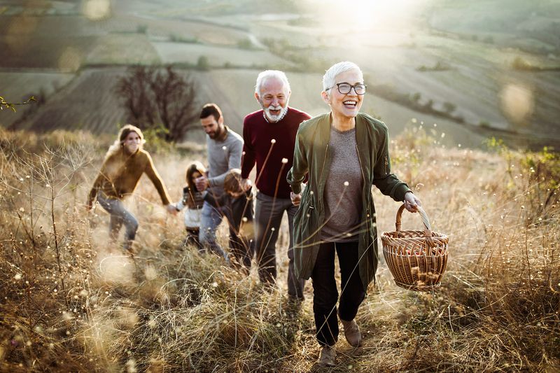 Grandparents and a young family with children laughs as they hold hands and climb a grassy hill to have a picnic.