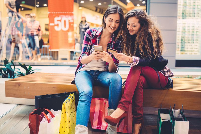 Two young women sit on a bench in a mall. 
