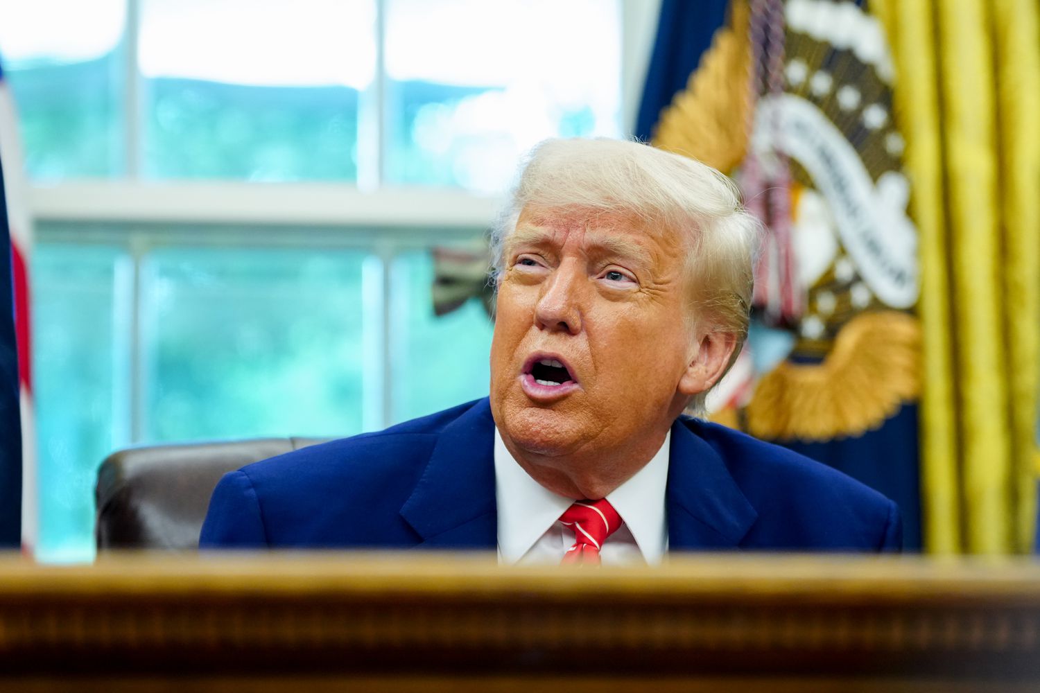 President Donald Trump sits behind a desk, with flags donning the presidential seal behind him. 