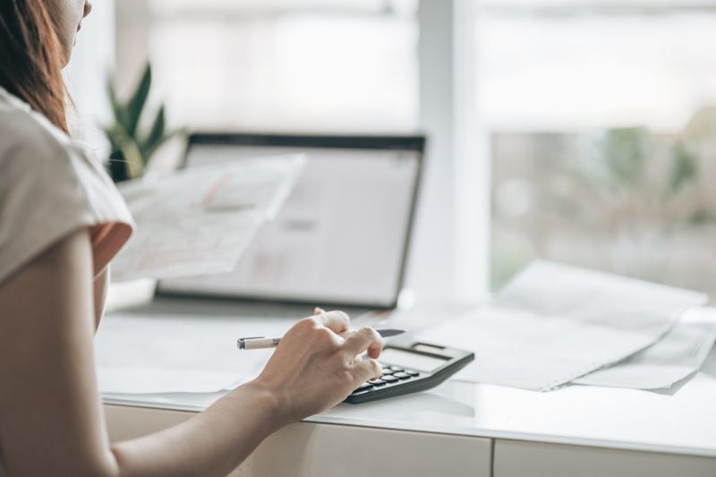Woman typing on calculator at desk while holding papers, and a computer in the background