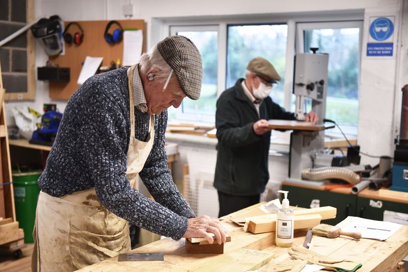 Two men work on woodworking projects in a carpentry workshop, one at a bench sanding, and the other in the background