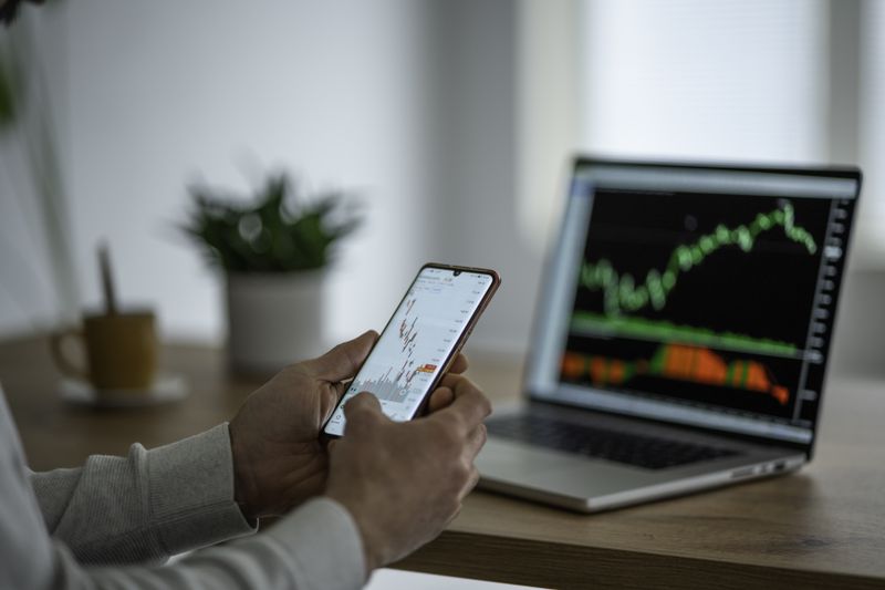 A person checks a stock market graph on his phone, with a laptop screen in the background
