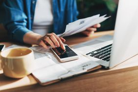 Close up of a person using a calculator and laptop for doing accounting on a wooden desk in an office.