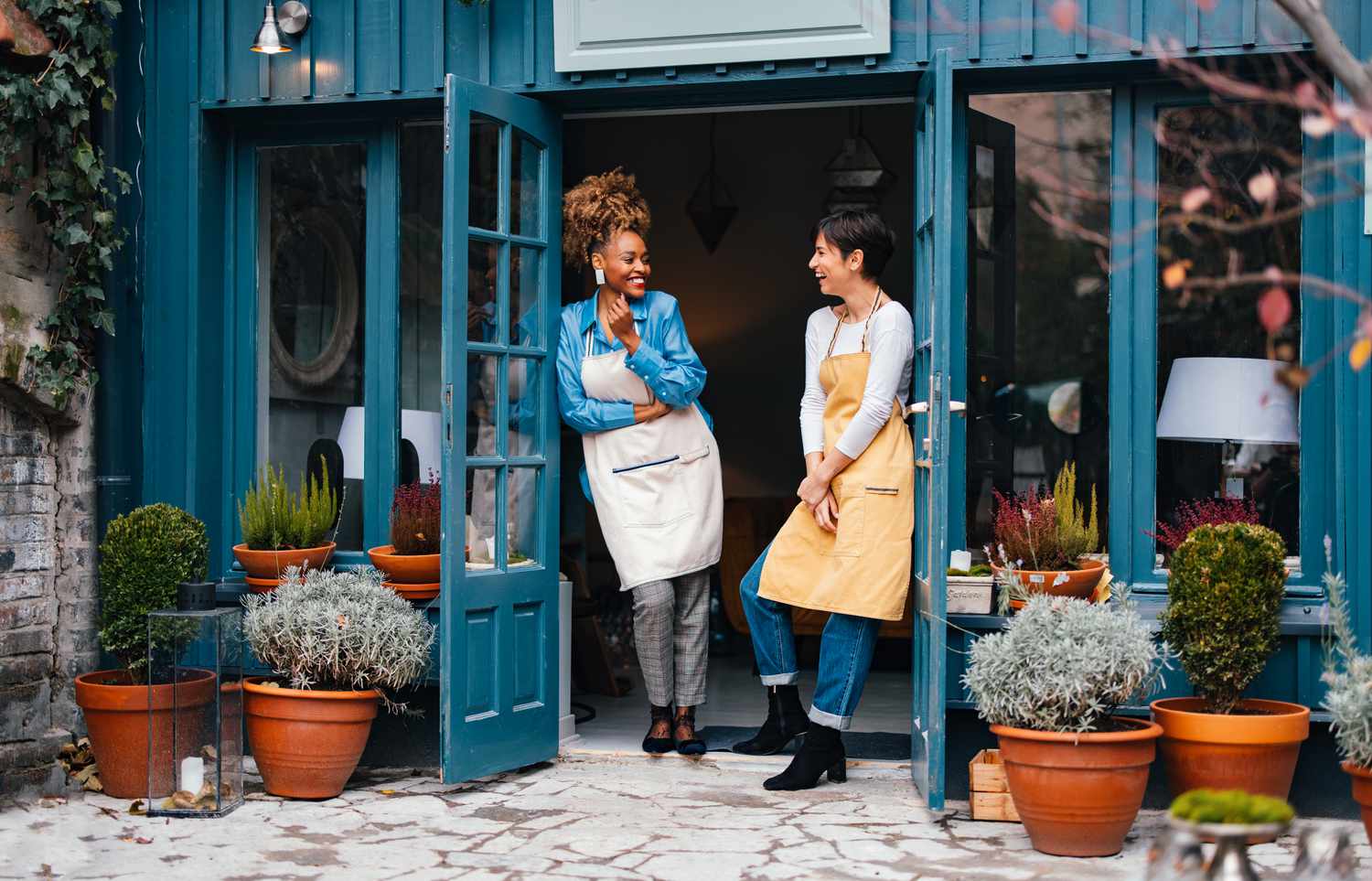 Two interior decorators and owners of a small business laugh as they share stories at the open doorway to their shop.