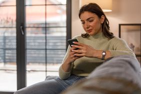 Young woman sitting in her living room and looking seriously at something on her smartphone