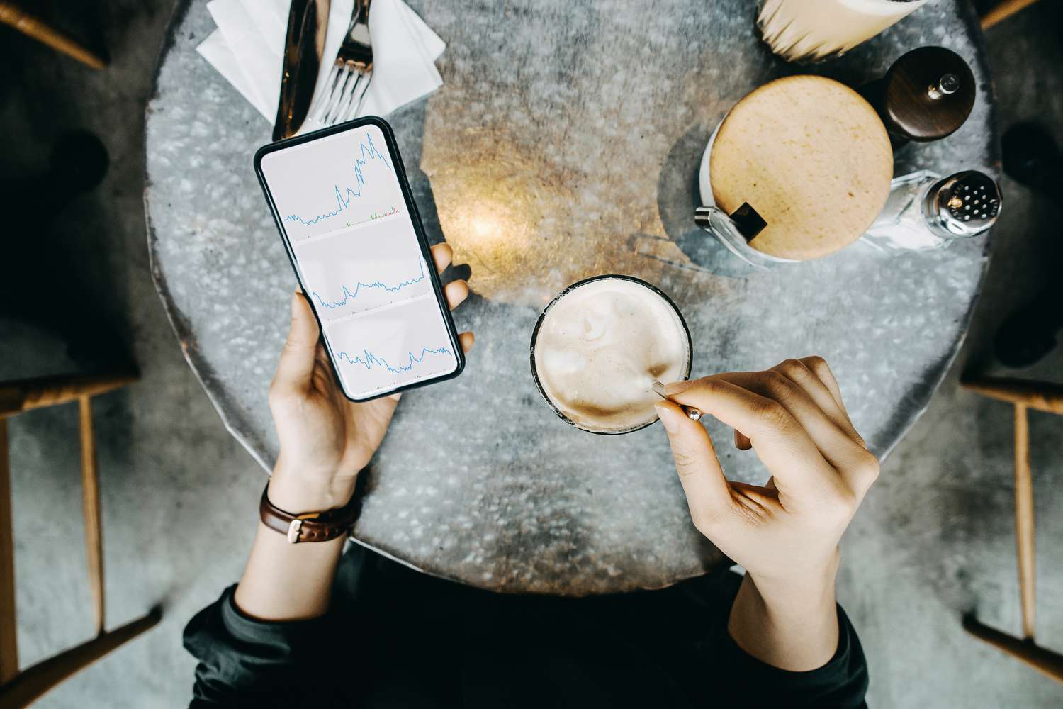 Overhead shot of a woman looking at her phone, which shows line graphs, while stirring a coffee