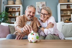 A happy couple deposits change into a piggy bank, seated on a couch.