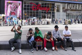 People sitting on a stone ledge in a city setting with a Disney store in the background, holding phones or in conversation