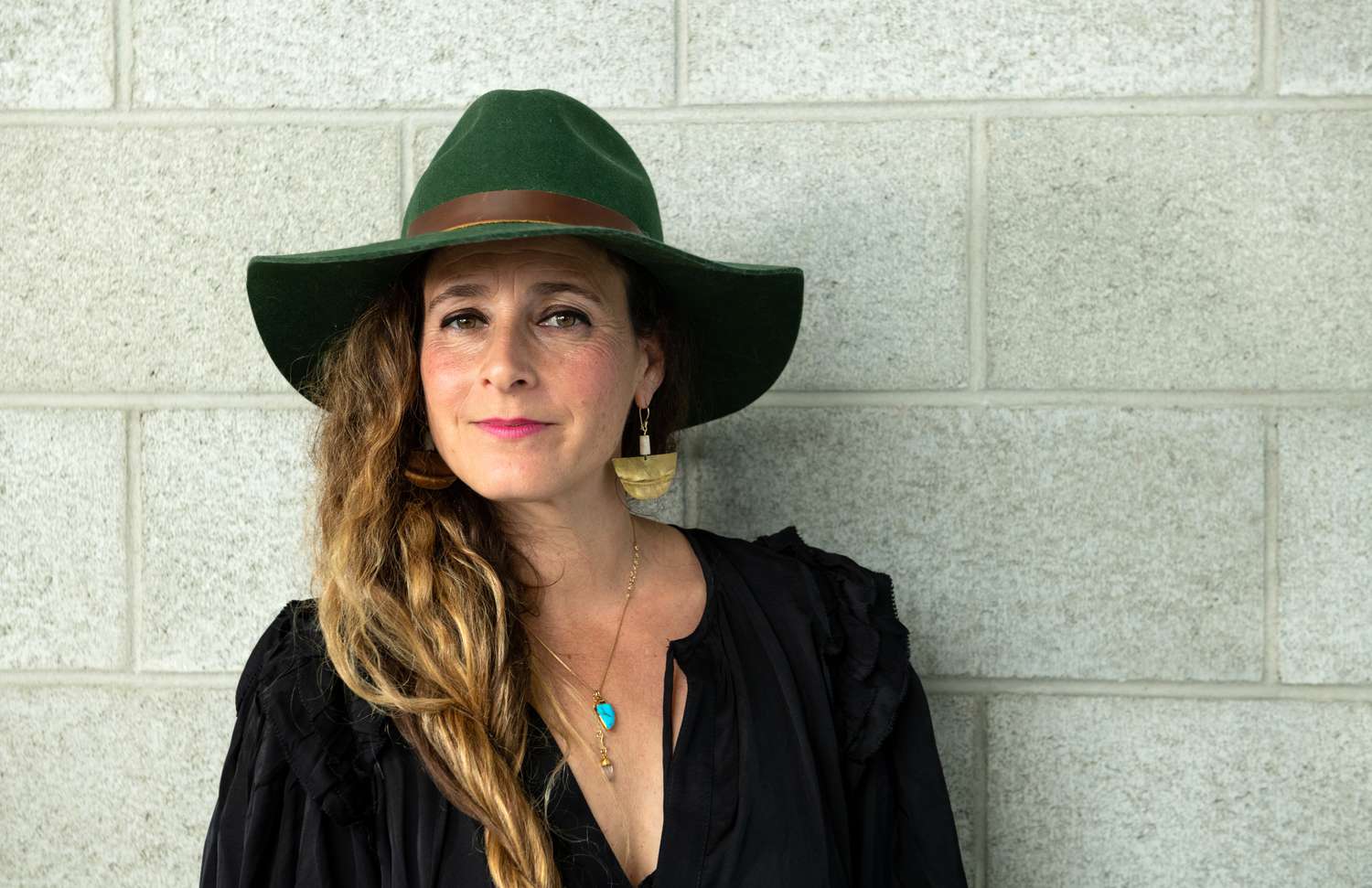A person wearing a wide-brimmed hat and earrings stands against a light background, looking towards the camera