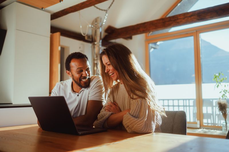 A couple is sitting together at a wooden table in their living room, engaged in a collaborative task on a laptop.