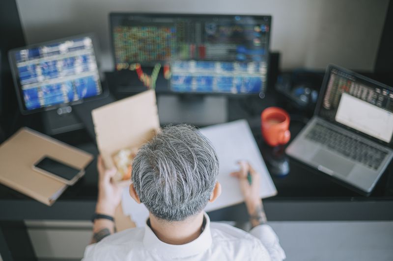 An image from above a gray haired man eating takeout food while working at home with a laptop and multiple screens