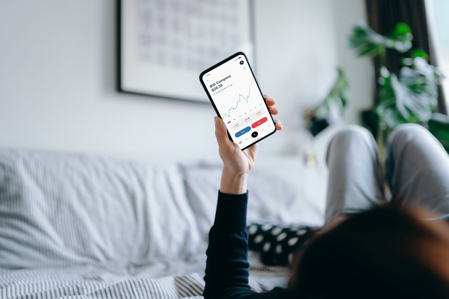 Young person lying on a bed in a home, reading financial data on a smartphone.