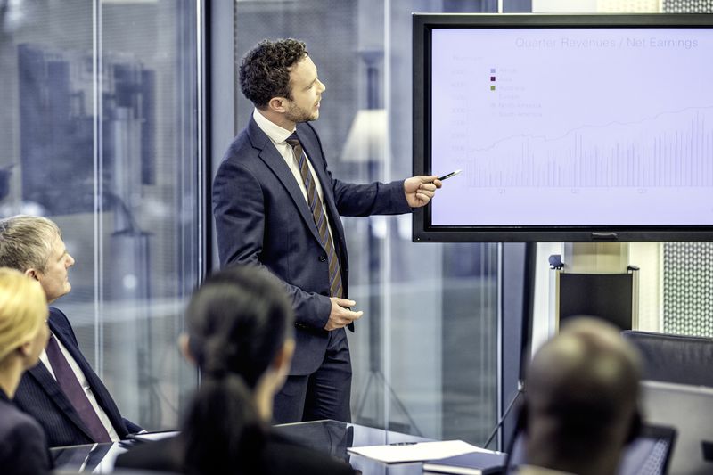 Financial analyst standing next to a monitor with graphs and explaining them to company board.