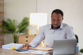 A man sits at a desk in front of a laptop and a calculator, reviewing papers that appear to display financial charts and data. 