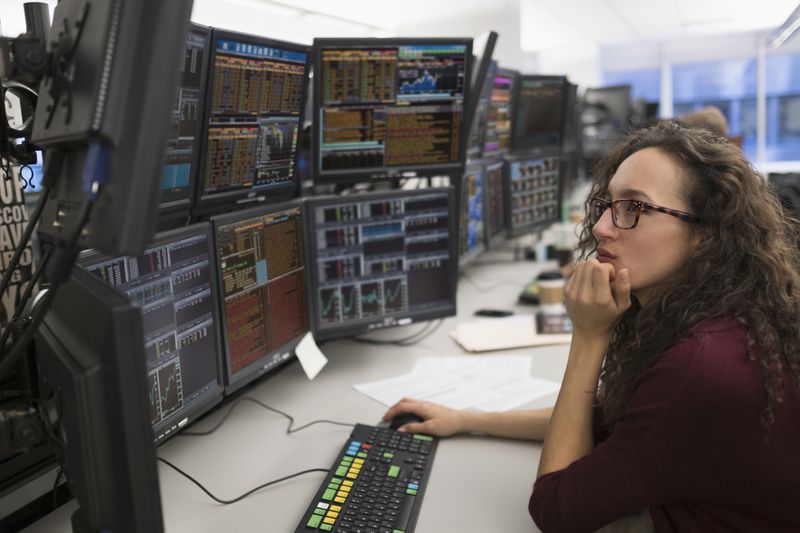 A young professional woman sits in front of multiple monitors displaying financial data.