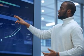 An analyst talks in front of a screen displaying quarterly results of a company they are recommending for an upgrade from hold to buy.