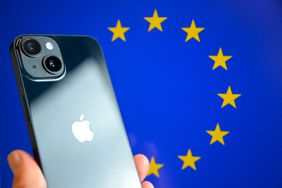 This photograph shows a hand-held smartphone with the Apple company logo and the European Union logo in the background