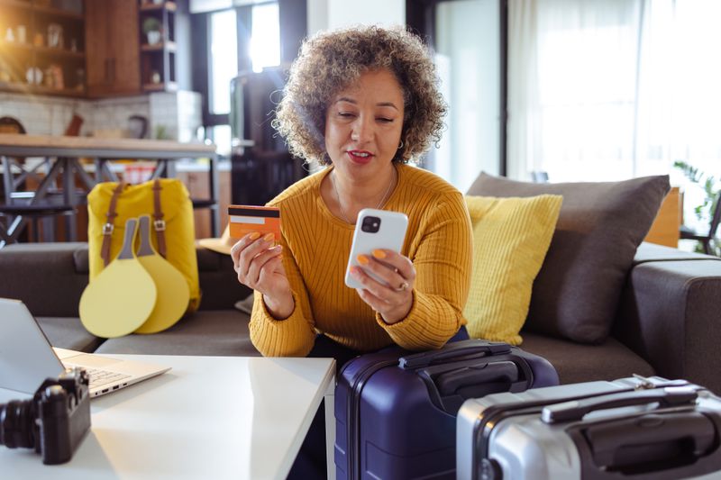 A woman in a yellow sweater sits in her living room with a credit card in her right hand while looking at a smartphone. She has two suitcases standing in front of her.