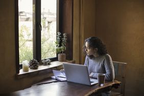 Young Woman of Mixed Ethnicity Works From Home Using Laptop Computer and Reference Book