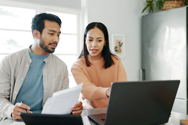 Young couple sitting in their kitchen and looking at mortgage documents and rates on a laptop