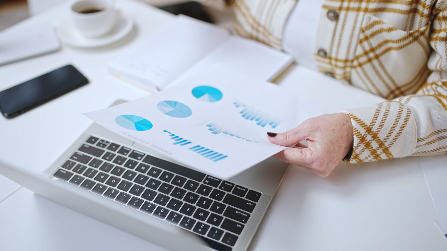 A woman sits at a desk with a laptop, holding a paper depicting financial charts and graphs.