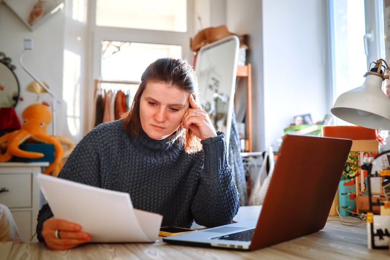 The image shows a woman wearing a gray knitted sweater sitting at a table in a cozy, well-lit room. She is holding a sheet of paper in one hand while resting her head on the other, appearing focused or deep in thought. In front of her is an open laptop. The background features a standing mirror, various personal items, and a colorful stuffed octopus. The setting suggests she is working or studying in a casual home environment.