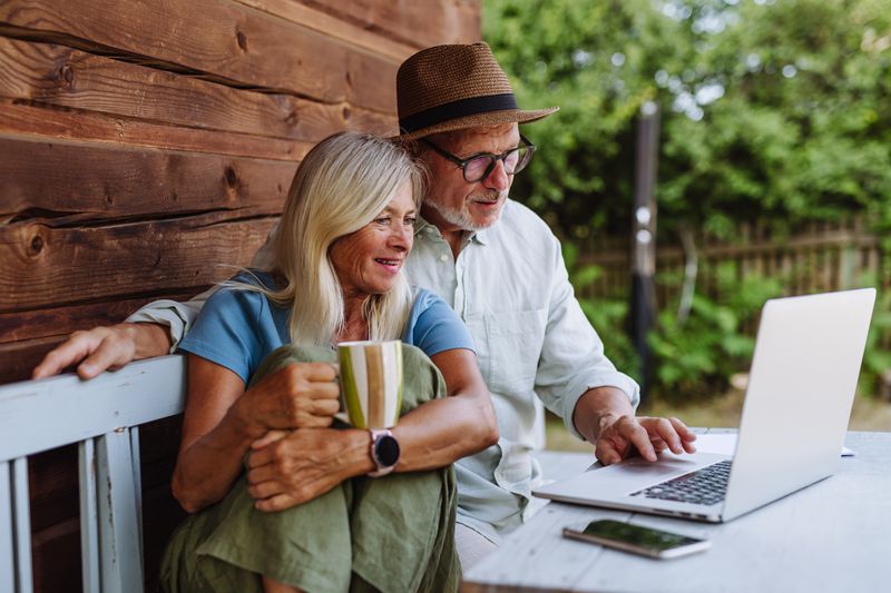 Two older adults sitting outdoors, one holding a mug, both looking at a laptop on the table