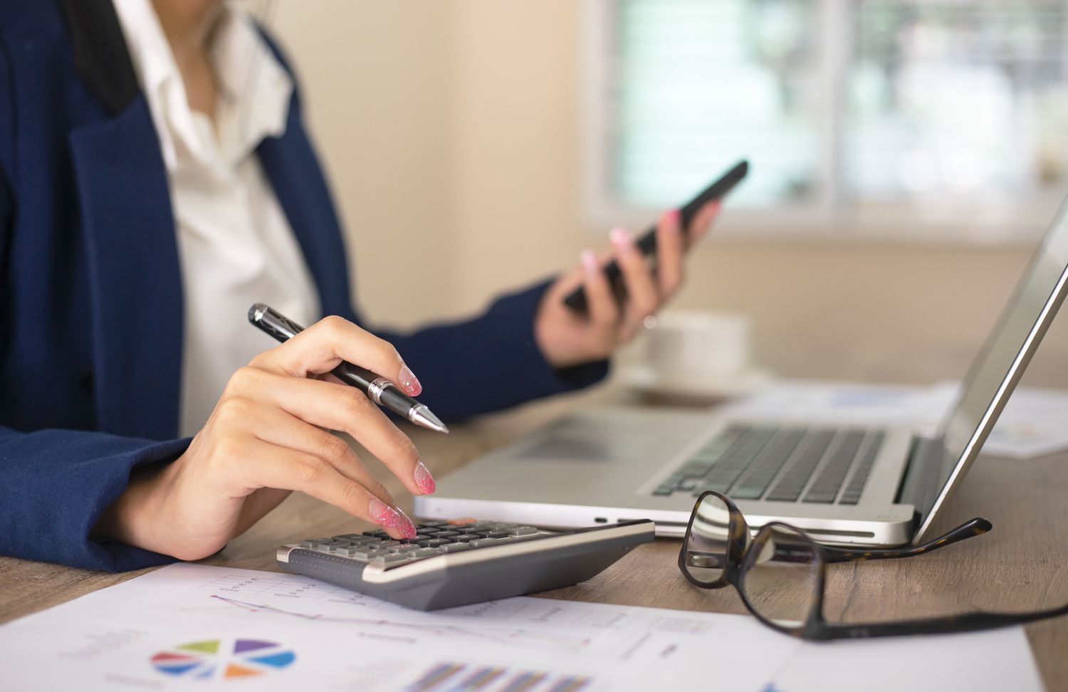 Businesswoman working in office typing on calculator with computer and papers on desk