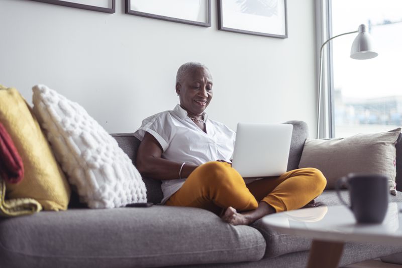 A black senior woman sits comfortably on her couch in her apartment. She is using a laptop.