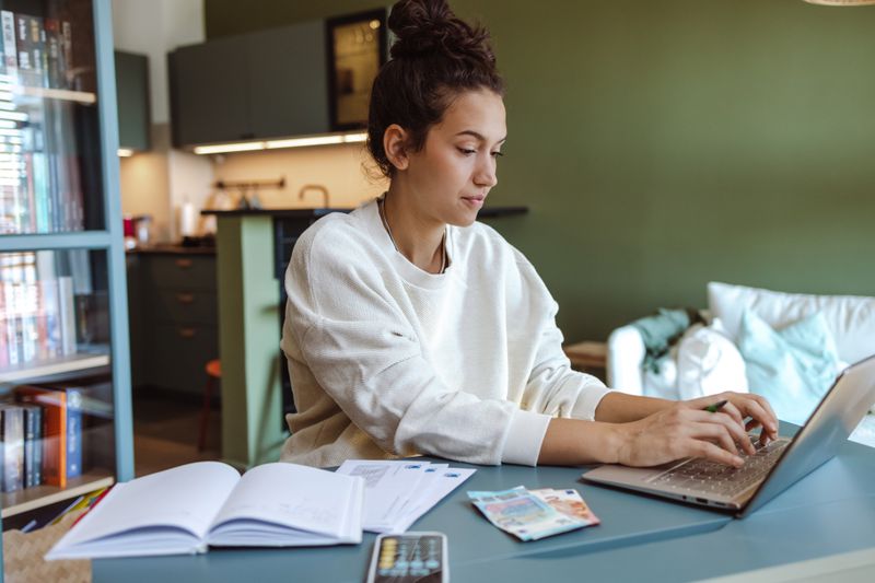 A woman sits at a table with a laptop and calculator, reviewing her budget 