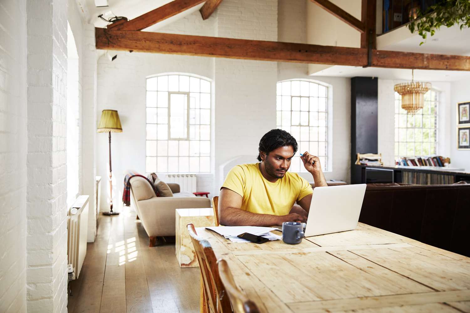A man in a yellow t-shirt sits at a wooden table at home with a laptop and mug