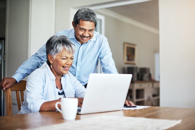 Two smiling adults look at a laptop on a kitchen table. 