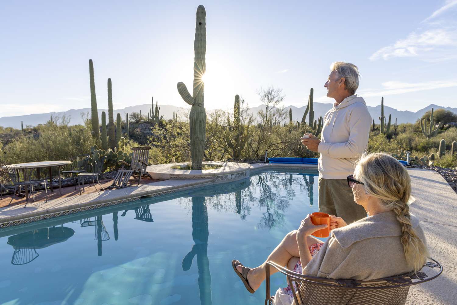 Two people holding mugs look out at the sunrise over a pool in Arizona. 