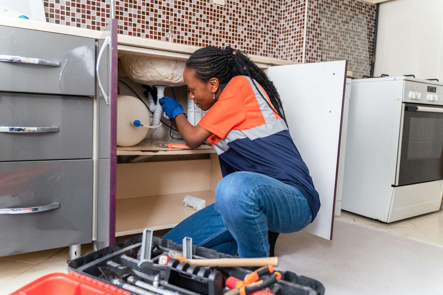 Black plumber in uniform sitting on the floor in kitchen and fixing pipe