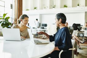 Two women sitting in an open plan work space, with laptops on the desk before them.