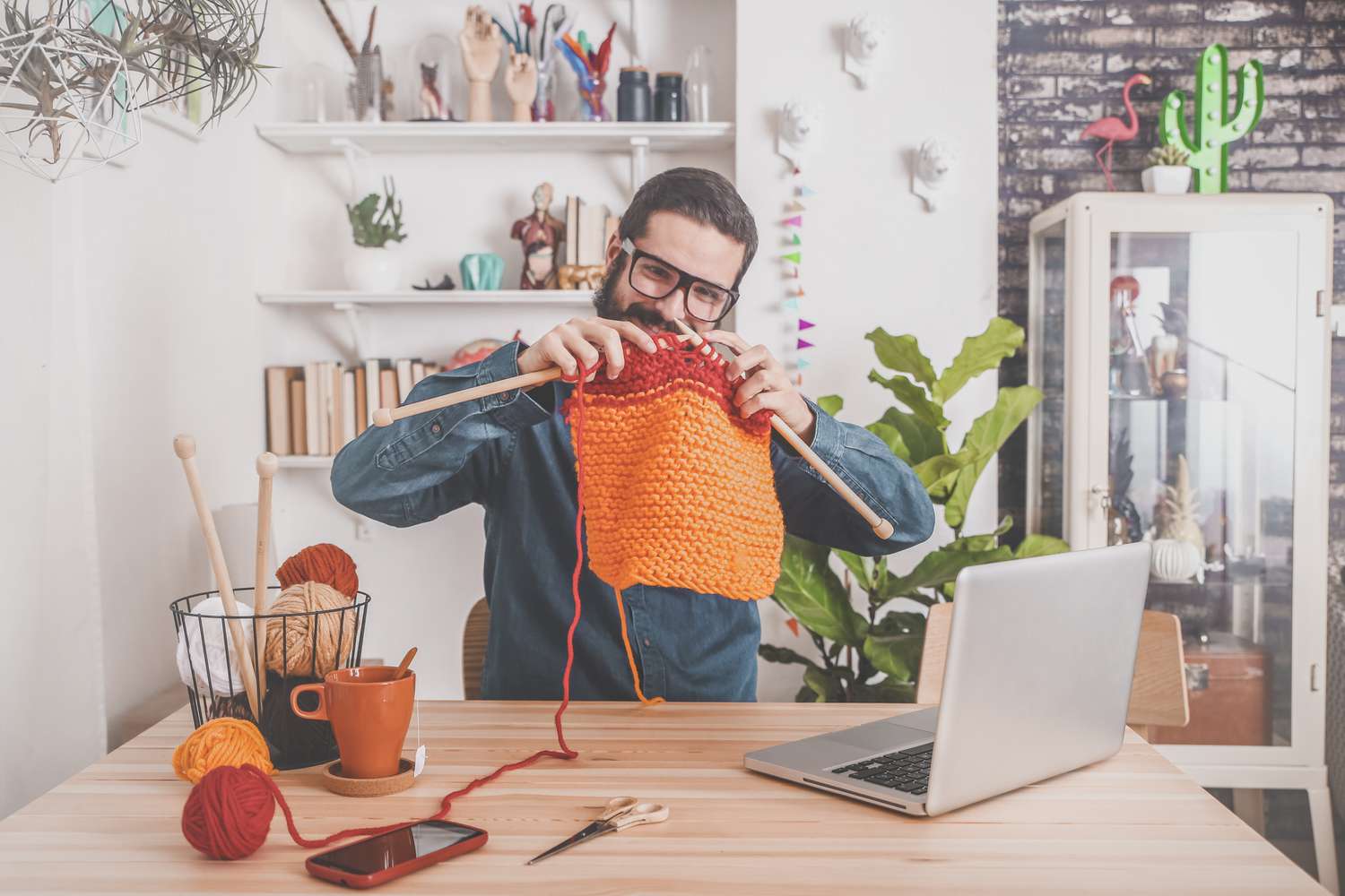 A person with a beard smiles while knitting at a desk in front of a laptop.