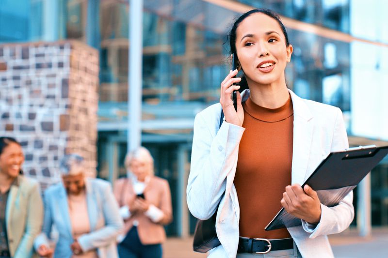  Businesswoman Talking On Phone With Colleagues In Background Outside Office Building