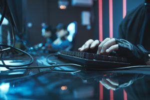 Close-up of someone's hands typing on a keyboard in an e-sports or gaming setup, blurred background shows players
