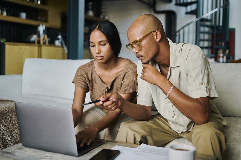 Two people sitting on a sofa, looking at a laptop together and holding a pen, appearing to discuss something