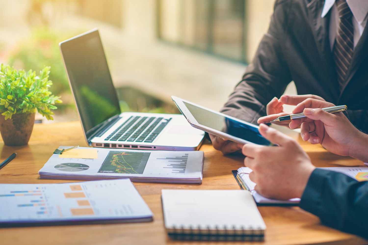 two people's arms and hands holding a tablet and a table with a laptop and finance reports on it