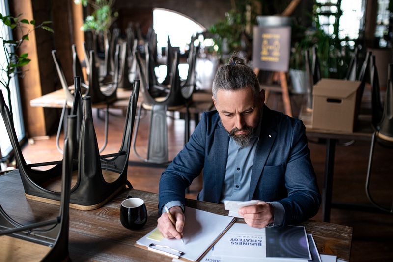 Small business owner calculating figures, looking at receipts in a business with stools turned upside down on tables